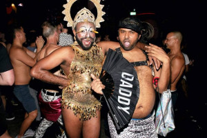 Two people in extravagant costumes pose for a photo. One wears a gold, fringed outfit with a headdress, and the other wears leopard print pants, a harness, and a cap, holding a black fan that says “DADDY.” The background shows a lively crowd dancing.