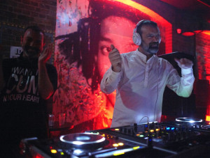 A DJ with headphones on stands behind a console, energetically playing music in a dimly lit room with red lighting. Another person stands beside him, clapping and smiling.