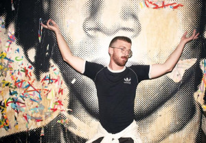 A man in a black Adidas shirt poses with his arms outstretched in front of a large mural featuring a stylized face. The mural has colorful, abstract paint splatters.