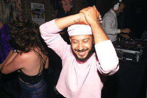 A man wearing a light pink outfit and head wrap dances with a big smile on his face. The background features other dancers and a DJ playing music on a set of turntables.