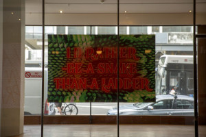 This image features a bold and colorful art installation displayed in a storefront window. The artwork includes the text I'D RATHER BE A SNAKE THAN A LADDER set against a backdrop that mimics the texture of green snake skin. The installation interacts with the urban environment, visible through the window’s reflection showing city life and passing vehicles.