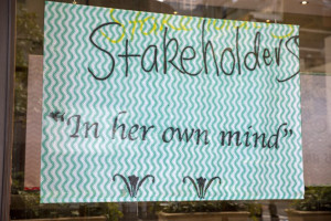 This image shows a handwritten sign in a window display, featuring the phrase Stakeholders In her own mind set against a zigzagging aqua and white background.