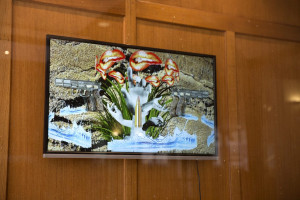 This image displays a flat screen mounted on a wooden panel showing a vivid and complex digital collage. The artwork features a juxtaposition of various elements including a pistol, vibrant red lips, and abstract forms that might suggest water and vegetation.