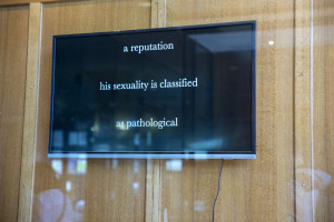 This image shows a flat screen display mounted on a wooden panel, featuring a text excerpt that reads a reputation his sexuality is classified as pathological. The text is presented in white on a dark background.