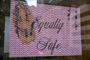 This image shows a sign displayed in a window with the message Equally Safe written in stylish script against a red and white chevron patterned background