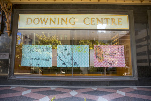 This image shows the glass facade of the Downing Centre, displaying colorful, patterned posters with handwritten messages. These posters are set against the backdrop of the building's interior, visible through the reflective window glass. The elegant, gold-trimmed lettering on the transom announces the name of the centre, framed by a traditional architectural detail, adding a classic touch to the otherwise modern view. The text on the posters suggests a possible art installation or public message display, contributing to the urban and cultural context of the location.