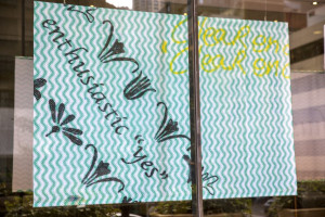 A large piece of fabric hangs in a shopfront window. Thee material features a vibrant zigzag pattern with the phrase 'enthusiastic yes' printed on top in black. Additionally, a whimsical floral pattern is also printed on top and below this phrase.