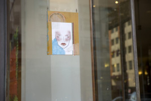 This image shows a piece of artwork suspended in a glass window display. The artwork features a face, with expressive eyes and delicate facial features drawn on a page of paper, which includes other sketch marks and handwritten notes. This setting in a public window invites passersby to view and reflect on the expressive, minimalist portrait.