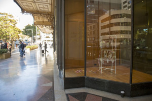 This image captures a reflective window display of an intricate sculpture placed inside a storefront. The artwork, possibly crafted from a clothes drying rack, features delicate, detailed components. Pedestrians can be seen walking along the sidewalk outside, reflected in the window, which blends the inside and outside environments.