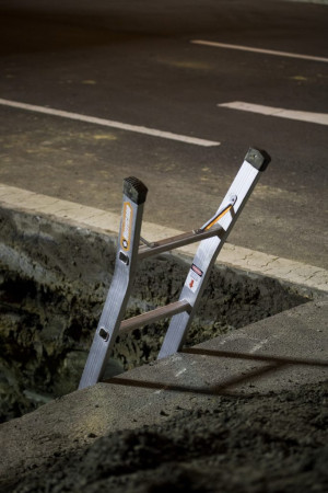 A metal ladder placed in a deep, rectangular excavation on a road, providing access to the underground area. The road surface is dark and the excavation edges are rough, with some gravel visible.