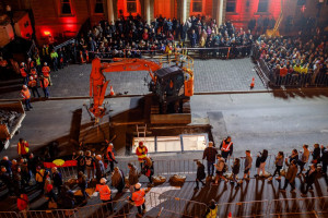 A nighttime scene of a construction site with a large crowd of people gathered behind barricades. Workers in orange safety vests are managing the site, where an excavator is positioned near an open underground structure. Bright lights illuminate the area, and the surrounding buildings are lit up with red lights.