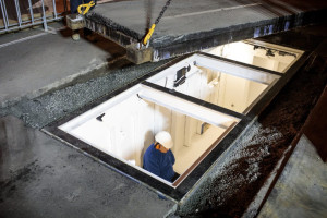 An artist in a white hard hat is seen from above through a glass ceiling, standing in a large, underground structure with white walls. The opening is surrounded by a concrete surface with gravel along the edges, and a metal frame outlines the window.