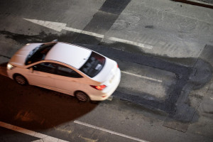 An overhead view of a white car driving on a road with a patchy surface. A large arrow is painted on the road, indicating the direction of traffic.