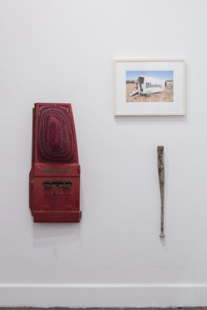 A wall-mounted art installation featuring a red object with a patterned fabric, a photograph of an old Mustang truck in a desert setting framed and hung on the wall, and a long, slender, weathered wooden object, resembling a club or bat.