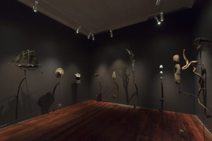 A dark-walled gallery room featuring various sculptures and installations. Objects include a miniature boat, coiled and woven items, and abstract forms mounted on thin metal stands, casting shadows on the walls.