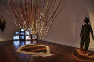 A dimly lit gallery room with multiple installations, including a cluster of vertical spears or sticks arranged like a burst, a stack of old televisions displaying various images, a patterned mat on the floor, and a wooden statue of a human figure standing on a tree stump.