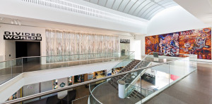 Modern art gallery space featuring the DIVIDED WORLDS exhibition. The gallery has a sleek, minimalist design with a curved ceiling and metallic railings. On the left, a translucent art installation hangs parallel to the wall, and on the right, a vibrant, large-scale abstract painting with intricate patterns dominates the view.