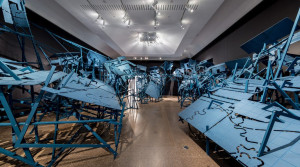 An art installation featuring an array of interlocking blue metal frameworks resembling geometric shapes and architectural components. The structures are arranged in a complex, sprawling formation across the gallery floor, creating an immersive and abstract environment. The installation is illuminated by ceiling lights, casting intricate shadows that contribute to the depth of the scene.