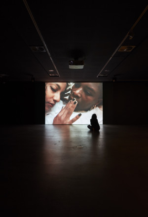 A person sits on the floor in a dark room, silhouetted against the large screen projection of a close-up image of two people's faces.