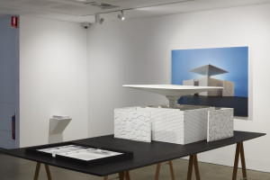 An art installation with a sleek, futuristic table model in a gallery setting, complemented by a large photographic representation of the model, evoking architectural design elements.