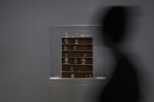 A glass display case containing a collection of small, sculpted heads arranged on wooden shelves. A blurred figure moves past the display in the foreground.
