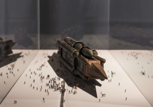 A detailed model of a large, futuristic vehicle surrounded by numerous small human figurines, displayed in a glass case.