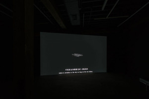 A dark room with a large video projection showing a small figure lying down. The subtitles on the screen read “which is invisible in the sky as long as the sun shines.”