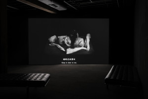 A dark room with a large black-and-white video projection showing a person interacting with large sculpted limbs. The subtitles on the screen read “Sleep is dear to me.” There are black cushioned benches in the foreground.