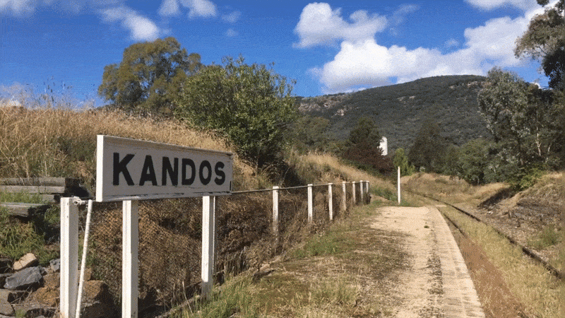 This image features a rustic scene with a dirt road passing by a sign that reads KANDOS. The road is flanked by a wooden fence and leads towards a lush mountain in the background under a clear blue sky, conveying a tranquil rural setting.