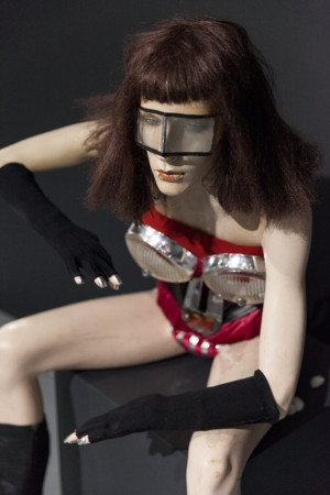  An art installation of a mannequin with a futuristic style. The mannequin has a reflective visor over its eyes, a red and silver bodice resembling armor, and is posed with one hand on its hip. It's wearing black gloves with metal tips and black boots, and is seated against a dark background.