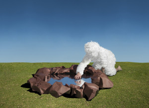 A conceptual art piece depicting a figure in a fluffy white suit that mimics the appearance of a cloud, bending over a hole with brown edges on a grassy field, against a clear blue sky. The hole is crafted to resemble a puddle or small pond, with the figure's reflection visible on the water's surface.