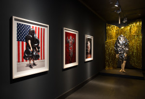 A gallery exhibition featuring a sequence of framed photographs on a dark wall, each showing a figure cloaked in unique, textured materials that obscure their identity. The first photograph depicts a figure wrapped in a glossy black material against an American flag, while the subsequent images explore different textures and backgrounds. A mannequin dressed in a reflective, metallic garment is positioned at the end of the wall, next to a golden tinsel curtain, extending the theme of the artworks into the three-dimensional space.