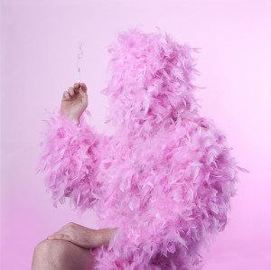 A creative and surreal image showing a figure covered entirely in pink feathers against a matching pink background. The person's head is not visible, and the only parts of the body that can be seen are an arm and a leg, revealing human skin. The figure's hand is holding a smoking cigarette.