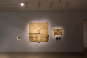 A gallery wall with three pieces of art, including woven mats and sculptures, lit by overhead lights.