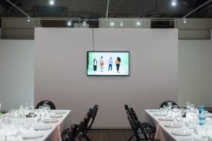 A dining space with elegant table settings in a row, and a flat-screen monitor on the wall displaying an image of four people in a green room.