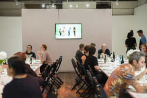 Diners at a gallery event with a lively atmosphere, seated at dining tables , while a video of dancers is displayed on a monitor in the background.