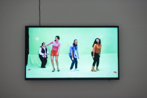 A video on a flat-screen display shows four individuals in various outfits performing a synchronized dance against a plain green backdrop.