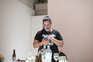 An individual in a black T-shirt reads from a smartphone into a microphone, standing at a dining event with tables set in front of them.