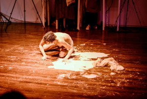 A shirtless performer crawls on a wooden floor covered with white liquid and fabric, with an audience watching from the background.