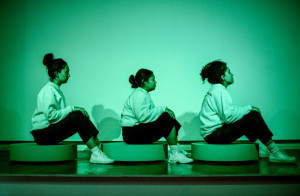 Three performers, each sitting on a circular platform, pose in identical seated positions, facing to the right, under green lighting. They wear matching white sweatshirts and dark pants.