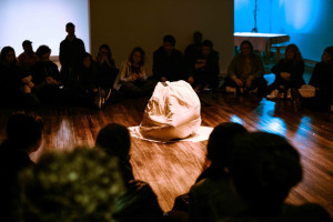 A group of people sit in a circle around a performer covered in white fabric in the center of a wooden floor, engaged in an intimate performance.