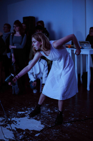 A performer in a white dress, holding a handheld light, leans forward and extends her arm during a performance. The audience watches in the dimly lit room with blue lighting.