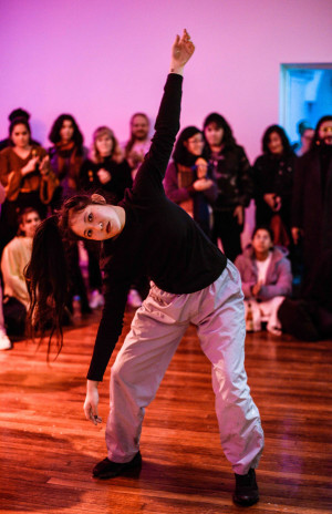 A performer with long dark hair, wearing a black top and light gray pants, strikes a dynamic pose with one arm raised and the other bent, in front of an audience watching attentively. The background is lit with a purple hue.