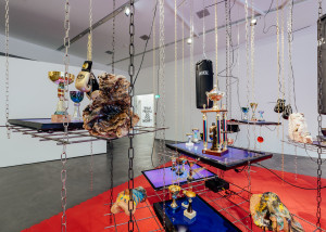 A contemporary art installation with assorted objects like trophies, a boxing glove, and a 'NIKU' punching bag, suspended by chains over a red mat in a white-walled gallery.