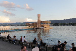 People relaxing on a lakeside promenade during dusk, with a unique wooden pavilion structure on the water and hills in the background.