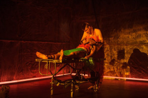 A dimly lit scene with a person lying on a medical gurney, partially covered in green and red fabric. Another person, with a shaved head and wearing a red apron, leans over the gurney, performing some action. The background is a metallic, textured surface illuminated with red lighting.
