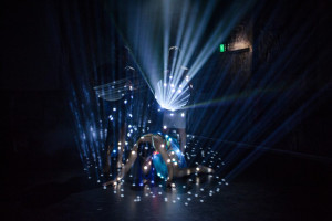 A performer on all fours is bathed in beams of light creating a pattern of dots around them. Two figures stand behind the performer, enhancing the dramatic effect with additional lighting and shadows.