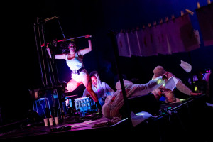 A performer in a white tank top and shorts energetically posing on stage, holding a pole above their head, with various objects and papers scattered around. Other performers are visible in the background, some dressed in white protective suits.