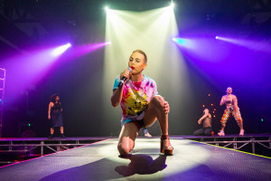 A person kneeling on stage, singing into a microphone, wearing a colorful tie-dye shirt and high heels. Other performers are dancing in the background under bright stage lights.