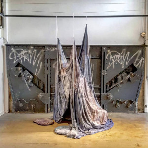 An industrial-themed art installation featuring distressed fabric suspended from the ceiling, creating a ghostly, draped form. The piece is set against a backdrop of a metal door with graffiti.